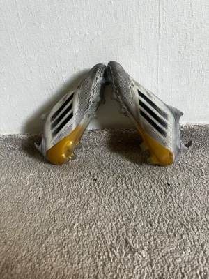 Vita och gula Adidas fotbollsskor - Säljer ett par snabba fotbollsskor från Adidas i vitt med svarta ränder och gula detaljer vid hälen. Skorna har låg profil och mönstrad ovandel för bra bollkontroll. Perfekta för dig som vill sticka ut på planen.