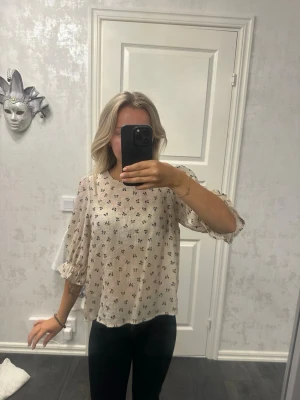 Beige blommig blus med trekvartsärm - Söt beige blus med små svarta blommor och trekvartsärmar med volangkant. Blusen har en lös och luftig passform, rund halsringning och är tillverkad i ett tunt, lätt material som ger en avslappnad look.