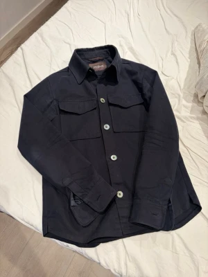 Mörkblå overshirt från Oscar Jacobson - Stilren mörkblå overshirt från Oscar Jacobson med klassisk krage, stora bröstfickor med lock och tydliga knappar framtill. Tillverkad i ett mjukt, slitstarkt tyg som ger en clean look. Perfekt lager-på-lager-plagg för dig som gillar minimalistisk stil.      Köpt för 3600 på Oscar Jacobsson i Gbg!!