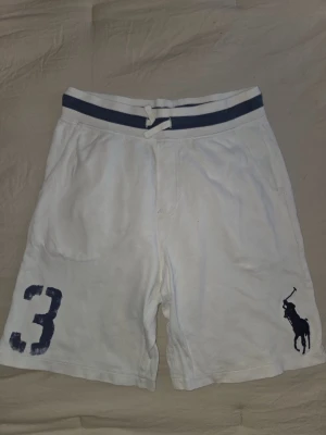 Vita shorts från Polo Ralph Lauren - Snygga vita shorts från Polo Ralph Lauren med blått tryck av siffran 3 på ena benet och den klassiska polospelaren på det andra. Shortsen har en blåvit randig resårmidja med dragsko och är tillverkade i mjukt bomullsmaterial. Perfekta för en avslappnad och sportig stil.