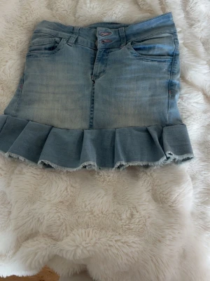 Ljusblå jeanskjol med volangkant - Trendig ljusblå jeanskjol med dubbla knappar och volangkant nedtill. Kjolen har klassiska fem fickor och råa kanter längst ner för en cool, avslappnad look. Perfekt att matcha med sneakers eller boots för en casual stil.