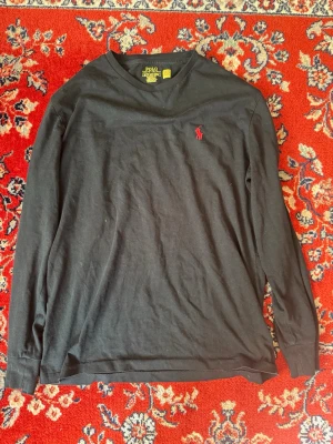 Svart långärmad tröja Polo Ralph Lauren - Snygg svart långärmad tröja från Polo Ralph Lauren i klassisk passform. Tröjan har rund halsringning och den ikoniska röda logotypen broderad på bröstet. Tillverkad i mjuk bomull som känns skön mot huden. Sitter som en s