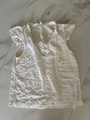 Vit broderad blus med knytband - Söt vit blus i broderad bomull med korta ärmar och volangkant vid halsen. Blusen har ett dekorativt knytband framtill och ett luftigt, avslappnat snitt. Perfekt för en somrig och romantisk look.