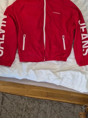 Röd vindjacka från Calvin Klein Jeans - Riktigt snygg röd vindjacka från Calvin Klein Jeans med vita dragkedjor och stora vita logotryck på ärmarna och ryggen. Jackan har två fickor med dragkedja och elastisk nederkant. Perfekt för en sportig och trendig look.