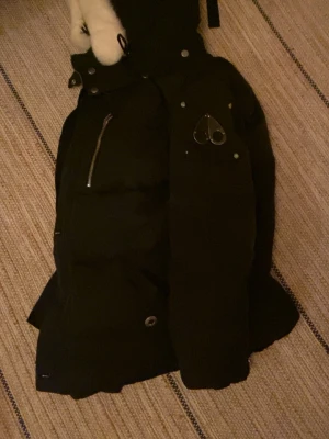 Moose knuckels jacka, storlek M - Snygg svart vinter jacka med flera silverfärgade dragkedjor och knappar. Jackan har en huva med vit fuskpäls och detaljer som en hjärtformad metallbricka på bröstet. Perfekt för kalla vinterdagar och riktigt varm.