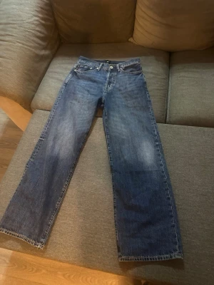 Blå raka jeans med normal passform - Säljer ett par klassiska blå jeans med raka ben och normal passform. Jeansen har fem fickor, knappgylf och är tillverkade i ett slitstarkt bomullsmaterial. Perfekta för en avslappnad och tidlös stil.