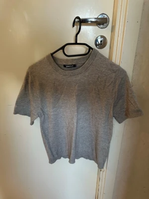 Grå kortärmad tröja från Weekday - En basic grå tröja från Weekday med rund halsringning och korta ärmar. Tillverkad i mjukt material som ger en clean och enkel look. Perfekt att styla med jeans eller kjol för en avslappnad vibe.
