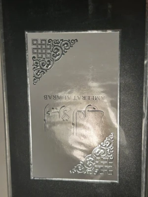 Amberrt Al Arab parfym - Snygg parfym med lyxig svart och silverfärgad förpackning. Designen har eleganta mönster i hörnen och text i både engelska och arabiska. Perfekt för dig som gillar exklusiva dofter och stilren design.