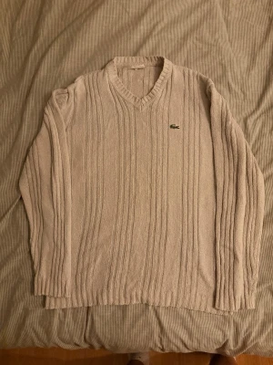 Beige stickad tröja från Lacoste - Säljer en beige stickad tröja från Lacoste med v-ringning och ribbade muddar. Tröjan har ett diskret Lacoste-märke på bröstet och ett snyggt kabelstickat mönster. Perfekt för dig som gillar klassisk och stilren design.