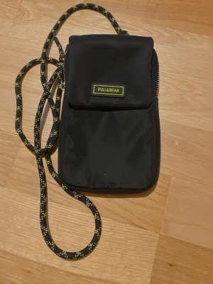 Svart telefonveska från Pull&Bear - Snygg svart telefonväska från Pull&Bear med justerbar snodd i svart och gult. Väskan har en praktisk ficka framtill med kapp och dragkedja på sidan. Perfekt för mobilen och småprylar. Tillverkad i slitstarkt tygmaterial.