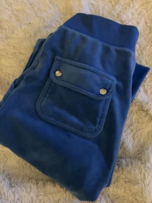 Juicy Couture byxor - Säljer ett par blå mjukisbyxor i mjukt velourmaterial. Byxorna har bred mudd i midjan med dragsko och stora fickor fram, samt en ficka med lock och tryckknappar bak. Perfekta för chill dagar eller när du vill vara extra bekväm.
