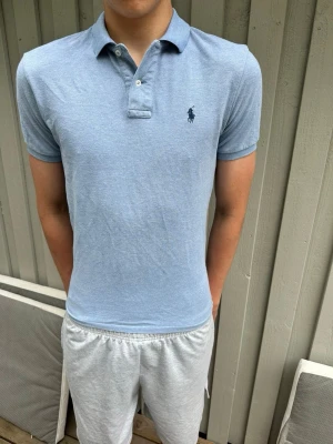 Ljusblå pikétröja från Ralph Lauren - Klassisk ljusblå pikétröja från Ralph Lauren med korta ärmar och krage. Tröjan har två knappar vid halsen och den ikoniska loggan broderad på bröstet. Perfekt till jeans eller shorts för en clean och stilren look.