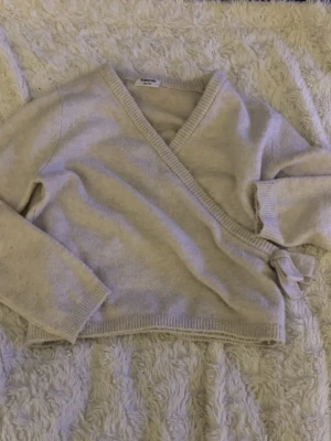 Beige omlottstickad tröja - Mysig beige stickad tröja med omlottmodell och knytband i sidan. Tröjan har långa ärmar och ribbade muddar vid ärmslut och nederkant. Perfekt för lager på lager och ger en avslappnad men ändå trendig look.