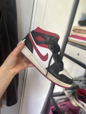 Svarta och röda Nike Air Jordan 1 Mid - Säljer ett par Nike Air Jordan 1 Mid sneakers i svart, vitt och rött. Skorna har klassisk swoosh-logga på sidan, svart läderdetalj, röd krage och yttersula samt vita paneler. Ikonisk Air Jordan-logga på sidan. Perfekta för dig som gillar streetwear. STORLEK 38