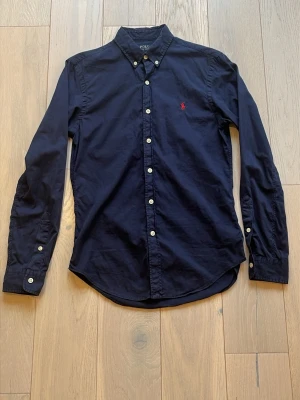 Mörkblå skjorta från Polo Ralph Lauren - Klassisk mörkblå skjorta från Polo Ralph Lauren med knappslå och vit knappdetalj. Skjortan har lång ärm och ikonisk röd logobrodyr på bröstet. Perfekt till jeans eller chinos för en clean och stilren look. Det är inga defekter eller annat men går alltid att höra av sig om man vill!👍