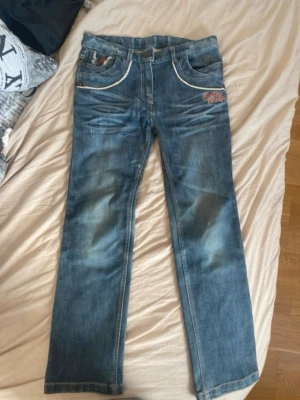 Blå jeans med vita sömmar och detaljer - Säljer ett par blå jeans med raka ben, vita kontrastsömmar och coola röda detaljer på fickorna. Jeansen har slitningar framtill och en broderad röd detalj på bakfickan. Klassisk femficksmodell i denim med knappstängning. osäker på storlek men skulle säga typ S 