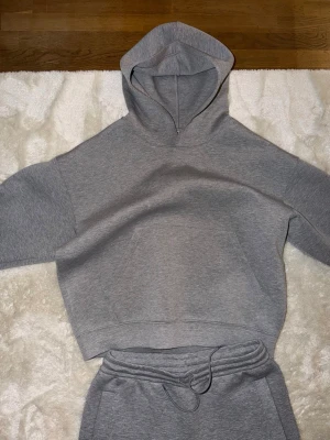 Grått scubaset med hoodie och byxor - Säljer ett grått mjukisset med hoodie och matchande byxor. Hoodien har en stor ficka framtill och byxorna har dragsko i midjan samt raka ben. Perfekt för chill dagar hemma eller när du vill vara bekväm. Materialet är mjukt och skönt mot huden.