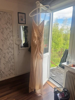 Glittrig champagnefärgad långklänning - Säljer en glittrig långklänning i champagnefärg med smala axelband och draperad v-ringning. Klänningen har en slits framtill och ett elegant fall. Perfekt för dig som vill sticka ut på bal eller fest. I storlek m priset kan diskuteras 