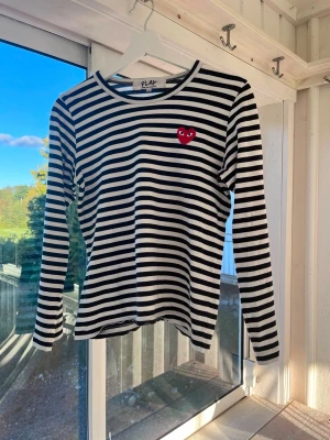 Randig långärmad topp Comme des Garçons - Säljer en svartvit randig långärmad topp från Comme des Garçons Play med det ikoniska röda hjärtat broderat på bröstet. Toppen har rund hals och är i mjuk bomull, perfekt för en stilren och trendig look. Passar dam S/M