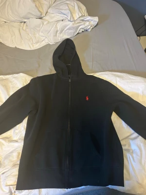 Svart hoodie med dragkedja från Polo Ralph Lauren - Säljer en svart hoodie med dragkedja från Polo Ralph Lauren. Klassisk design med huva och fickor framtill. Den har det ikoniska röda Polo-logotypen broderad på bröstet. Perfekt för en chill och stilren look. Pris kan diskuteras!