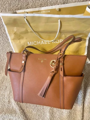 Brun handväska från Michael Kors - Snygg brun handväska i läder från Michael Kors med guldiga detaljer och tofs. Väskan har två handtag, axelrem, flera fack och dragkedja. Perfekt för dig som vill ha plats för allt du behöver. Klassisk och stilren design. Ordinare pris är för 4200kr.
