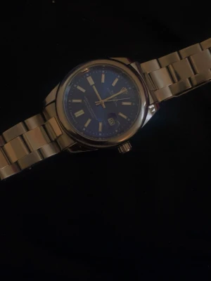 Klassisk silverfärgad armbandsklocka - Stilren armbandsklocka i rostfritt stål med blå urtavla och datumvisning. Klockan har tydliga indexmarkeringar och en länkad metallrem som ger en tidlös look. Perfekt accessoar för dig som gillar klassisk och enkel design.