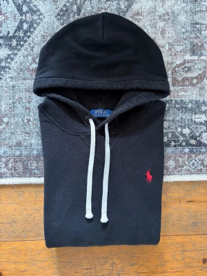 Svart hoodie från Polo Ralph Lauren - Klassisk svart hoodie från Polo Ralph Lauren med vit dragsko och röd broderad logga på bröstet. Hoodien har känguruficka framtill och ribbade muddar vid ärmar och nederkant. Perfekt för en avslappnad och stilren look. Personen på bilden är ca 177cm lång. Tröjan upplevs mer som en xs i längden av tröjan. Rekommenderad längd ~170cm