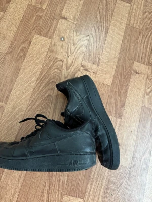 Svarta Nike Air Force 1 sneakers - Klassiska helsvarta Nike Air Force 1 sneakers i läder med låg profil och svart sula. Skorna har den ikoniska swoosh-loggan på sidan och AIR-detalj på sulan. Perfekta för en streetwear-look och passar till det mesta.