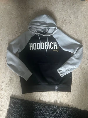Svart och grå HOODRICH hoodie - Snygg hoodie från HOODRICH med svart kropp och ljusgrå ärmar och huva. Stor vit logga framtill och tryck i gult och vitt på ryggen. Klassisk känguruficka och dragsko i huvan. Perfekt för en streetwear-look.