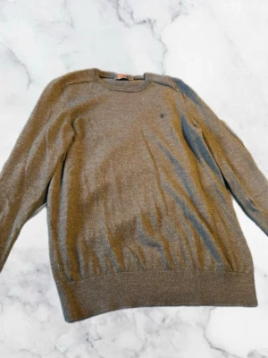 Brun stickad tröja från Morris - Säljer en stilren grå stickad tröja från Morris. Tröjan har rund halsringning, diskret logga broderad på bröstet och ribbade muddar vid ärmslut och nederkant. Perfekt att matcha med jeans eller chinos för en clean look. Tyvärr ett hål som syns i bilderna men inget som märks värst mycket vid användning.