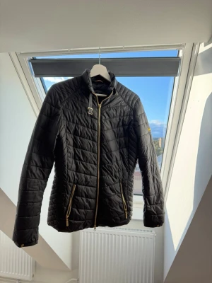 Svart quiltad pufferjacka med guldzip - Snygg svart pufferjacka med quiltat mönster och guldfärgade dragkedjor. Jackan har hög krage med spänne, två fickor med zip och en smal, figurnära passform. Perfekt för kalla vinterdagar och ger en stilren look.
