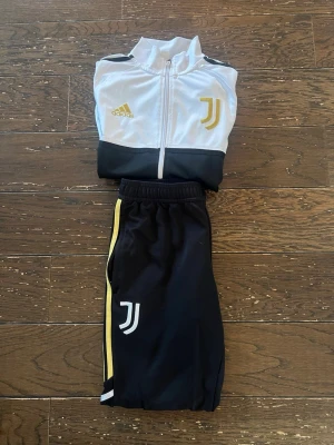 Juventus Adidas träningsset - Juventus träningsset från Adidas med vit och svart zip-jacka och matchande svarta byxor. Jackan har guldiga detaljer, Juventus-logga och Adidas-märke, samt svarta ränder på ärmarna. Byxorna har gul sidolinje och Juventus-logga. Perfekt för fotbollsträning. Tröjan har en liten fläck på ryggen. Priset är inte hugget i sten