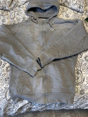 Grå zip hoodie från Gymshark - Säljer en grå hoodie från Gymshark med dragkedja framtill och justerbar huva. Hoodien har lång ärm, ribbade muddar och en liten vit Gymshark-logga på bröstet. Perfekt för chill eller gymmet.