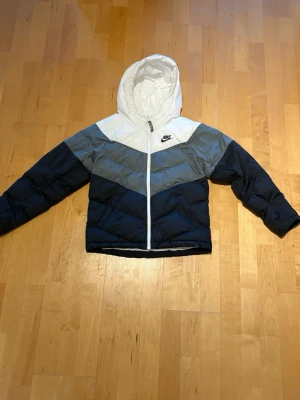 Nike pufferjacka med huva - Snygg pufferjacka från Nike i färgblock med vitt, grått och mörkblått. Jackan har huva, dragkedja framtill och klassisk Nike-logga på bröstet. Perfekt för kalla dagar och riktigt skön att bära. Materialet är vadderat syntet för extra värme.