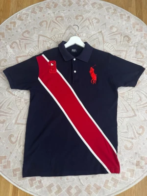 Polo Ralph Lauren Pikétröja - Storlek: M Färg: Marinblå/Röd Skick: Mycket bra  Klassisk pikétröja från Polo Ralph Lauren med stort broderat polospelaremblem framtill och röd/vit detalj över bröstet. Stilren modell med krage.  Se bilder för skick och detaljer.
