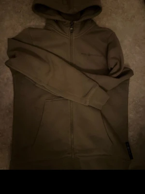 Olivgrön hoodie från Sail Racing - En helt ny snygg olivgrön hoodie från Sail Racing med dragkedja framtill och stor känguruficka. Hoodien har en broderad logga på bröstet och en klassisk huva. Tillverkad i mjukt material som känns skönt mot huden. Perfekt för chill dagar eller när du vill ha en clean look. pris går o diskutera (fraktar inom 8h 