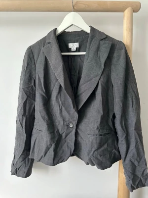Mörkgrå klassisk kavaj - Stilren mörkgrå kavaj med figurnära passform och klassisk krage. Kavajen har två framfickor, enkelknäppning med en knapp och dekorativa knappar vid ärmsluten. Tillverkad i ett tunt, vävt material som ger en dressad look. USA storlek 12, motsvarar runt storlek L.  Har förvarats i förråd. Kommer att skickas i nytvättad skick.