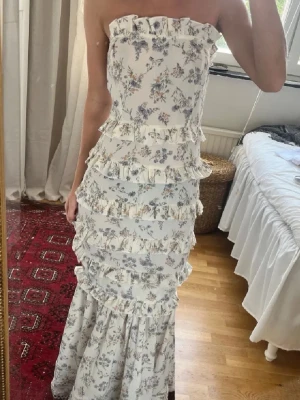 Blommig långklänning med volanger -  axelbandslös långklänning i vitt med blommigt mönster i lila och grå toner. Klänningen har flera volangdetaljer längs hela plagget och en figurnära passform. Från boohoo