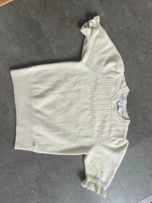 Vit stickad topp Holly & Whyte - Söt vit stickad topp från Holly & Whyte med kort puffärm och ribbade kanter. Ovanför bröstet finns ett dekorativt hålmönster som ger en romantisk känsla. Perfekt till jeans eller kjol för en trendig look. 