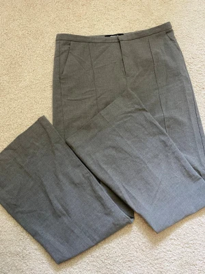 Bootcut Grå kostymbyxor med pressveck - Snygga grå kostymbyxor med raka ben och klassiska pressveck framtill. Byxorna har diskreta sidofickor och är tillverkade i ett tunt, vävt material som ger en stilren look. Perfekta för dig som vill ha en clean och tidlös stil.