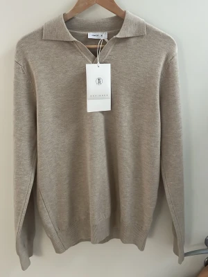 Beige stickad tröja med krage - Stilren beige stickad tröja från COOPER med klassisk krage och v-ringning. Tröjan har långa ärmar och ribbade muddar vid ärmslut och nederkant. Perfekt för en avslappnad men snygg look.