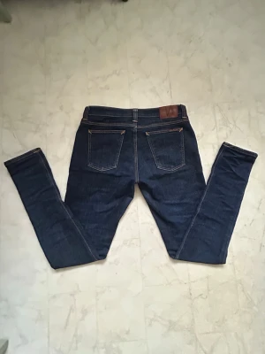 Mörkblå jeans från Nudie - Mörkblå jeans från Nudie med klassiska bakfickor, kontrastsömmar och läderpatch i midjan. Snygg passform och stilren design, perfekta till sneakers eller grisch stilen.  