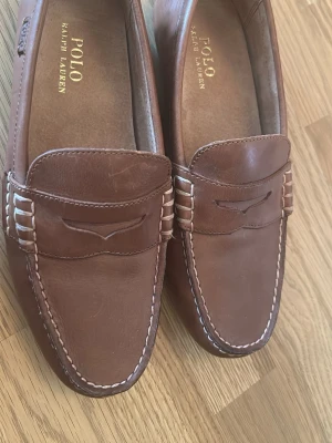 Bruna loafers från Polo Ralph Lauren - Snygga bruna loafers i läder från Polo Ralph Lauren. Skorna har klassisk design med dekorativa sömmar och detaljer på sidan samt gummisula för extra grepp. Perfekta för en stilren och avslappnad look.