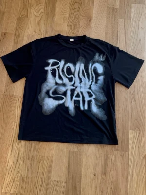 Svart t-shirt med sprayad text - Cool svart t-shirt med vit sprayad text på framsidan där det står 'RISING STAR'. Perfekt för dig som gillar streetwear och vill sticka ut med en unik design. Och storleken är 13-14 teen/XS.