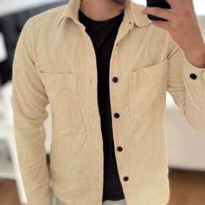 Beige overshirt i manchester - Snygg beige overshirt i manchestertyg med svarta knappar och två bröstfickor. Skjortan har klassisk krage och raka ärmar, perfekt att bära öppen över en t-shirt. Materialet är mjukt och ger en avslappnad men stilren look.