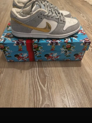 Nike SB Dunk Low Sean Cliver - Nike SB Dunk Low Pro QS sneakers i grått och vitt med en metallic guld swoosh. Skorna har klassisk låg siluett, perforerad tåbox och är tillverkade i mocka och läder. Snygg detalj med färgglad specialbox. Perfekta för dig som gillar streetwear och unika sneakers.