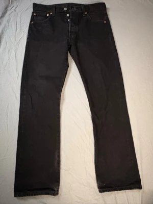 Levis 501 - Säljer ett par svarta Levis 501 jeans med rak passform och knappgylf. Jeansen har klassisk femficksdesign och detaljer i metall. Materialet är kraftig denim som ger en tidlös look.