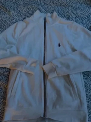 Vit zip hoodie från Polo Ralph Lauren - Säljer en vit hoodie från Polo Ralph Lauren med dragkedja framtill och två fickor. Klassisk broderad logga på bröstet och hög krage. Tillverkad i mjukt bomullsmaterial, perfekt för en clean och stilren look.