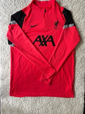 Nike Liverpool träningströja - Snygg röd långärmad träningströja från Nike med Liverpool FC-logga och AXA-tryck på bröstet. Tröjan har svarta detaljer på axlarna, halv dragkedja framtill och är tillverkad i ett lätt och funktionellt material som passar perfekt till fotbollsträning.
