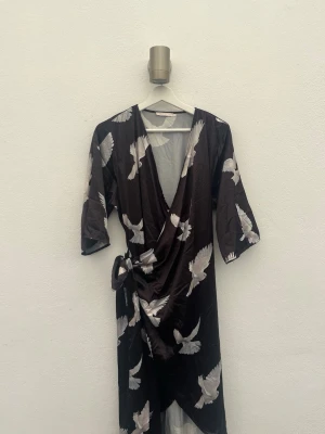 Svart kimono klänning med fågelmönster - Snygg svart morgonrock i kimonomodell med vitt fågelmönster. Lång och luftig med trekvartsärmar och knytband i midjan. Tillverkad i ett glansigt, silkesliknande material som känns lyxigt och bekvämt. 